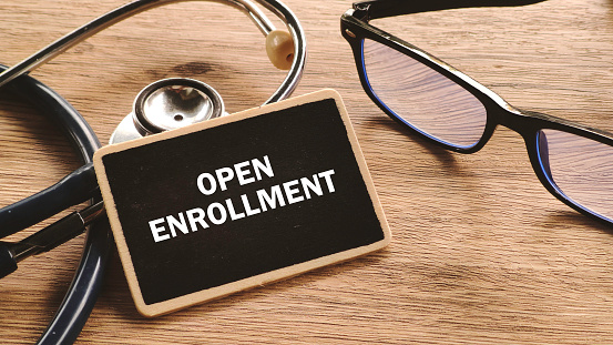 Top-down view of a wooden desk with a stethoscope, glasses, and a small chalkboard displaying OPEN ENROLLMENT.